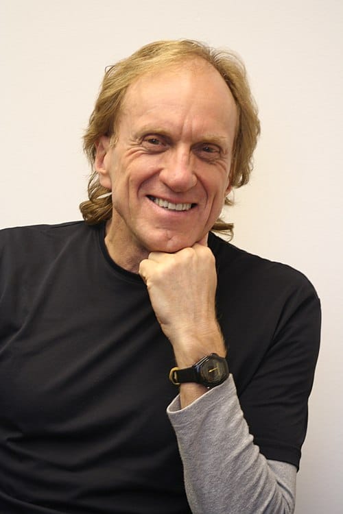 Smiling man in black shirt, resting chin on hand.